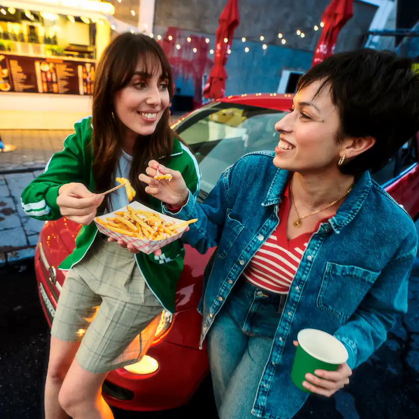 Two women eating fries together in front of a red car