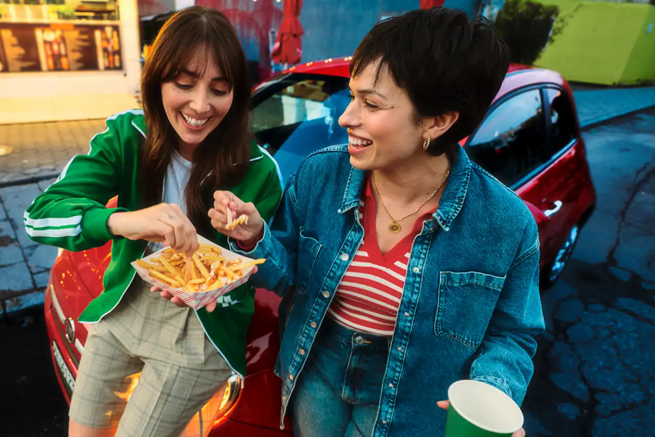 Two women eating and drinking sitting on a car